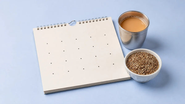 Spiral paper calendar with uneven dots beside a steel cup of chai and a bowl of millets