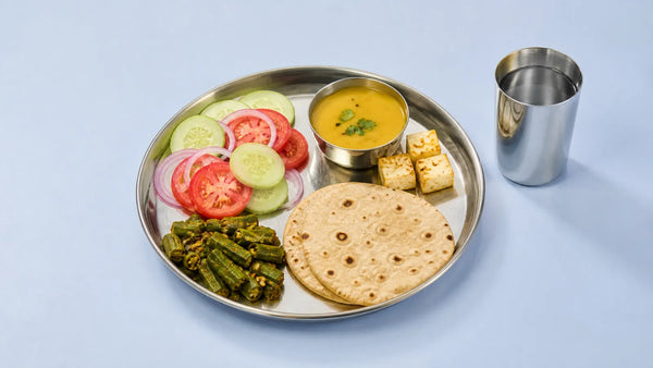 Steel thali with salad, bhindi sabzi, dal, paneer cubes and two rotis, with a steel glass of water