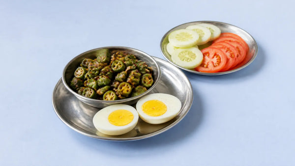 Two halves of a boiled egg on a steel plate beside a katori of bhindi sabzi and a plate of cucumber and tomato
