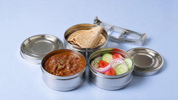 Three open steel tiffin boxes side by side with rotis, rajma and a cucumber, tomato and onion salad, lids beside them
