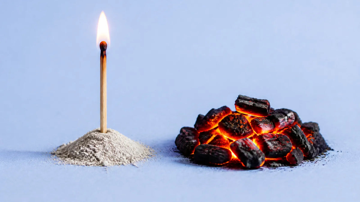 A lit matchstick standing in a small mound of ash beside a low pile of glowing charcoal embers on a pale blue surface