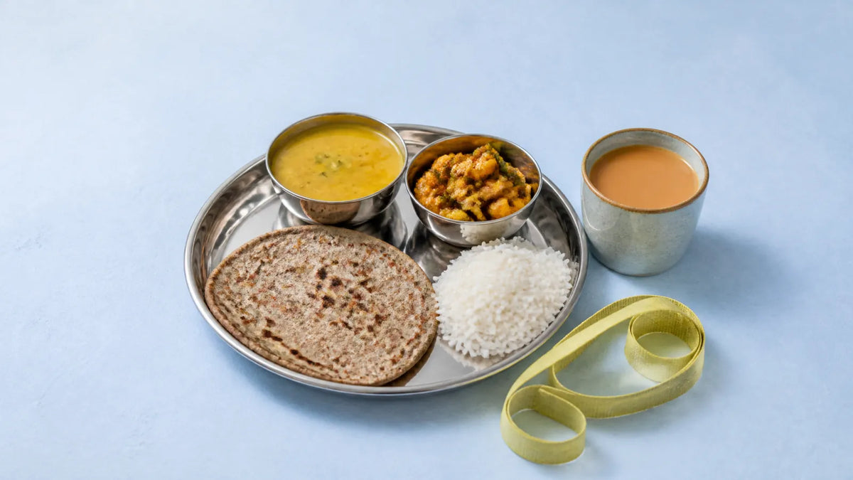 A steel thali with a millet roti, katoris of dal and sabzi, a little rice and a cup of chai beside a coiled cloth tape