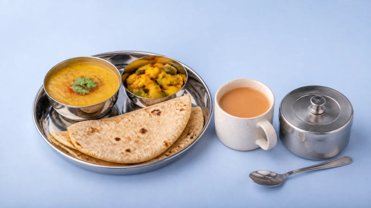A steel thali with rotis, dal and sabzi, a cup of chai and a closed steel sugar bowl with a spoon beside it