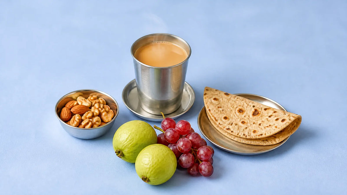 Steel tumbler of chai on a saucer beside a katori of almonds and walnuts, a folded chapati, two guavas and red grapes