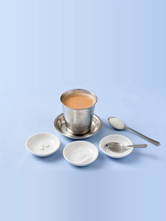 A steel tumbler of chai with dishes of sweetener tablets, a torn sachet and drops on a spoon, beside a spoon of sugar