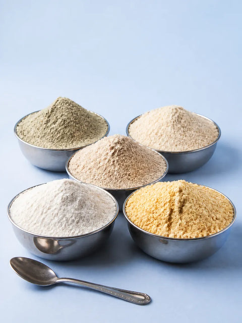 Five steel katoris heaped with maida, besan, atta, bajra and jowar flour on a pale blue table