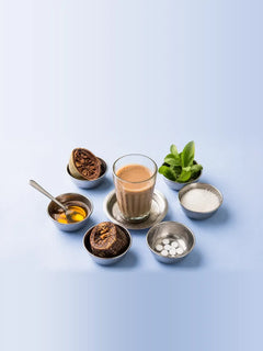 A glass of masala chai ringed by six steel katoris of monk fruit, stevia, white granules, tablets, gur and honey on a pale blue table