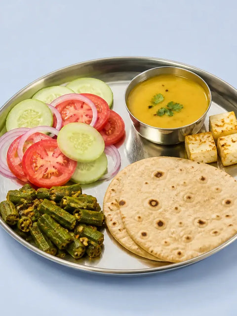 हल्की नीली मेज़ पर स्टील की थाली में सलाद, भिंडी की सब्ज़ी, दाल, पनीर के टुकड़े और दो रोटी