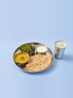 A steel thali with dal, a green sabzi, two rotis and a bowl of dahi, with a steel glass of chaas beside it on a pale blue table