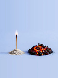 A burning match standing in a mound of ash beside a glowing pile of embers on a pale blue background
