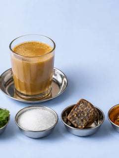 हल्की नीली मेज़ पर स्टील की तश्तरी पर चाय का गिलास, नीचे कटोरियों में मोंक फ्रूट, स्टेविया की पत्तियां, सफ़ेद चीनी, गुड़ और शहद