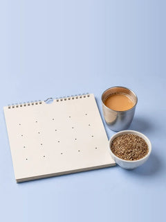A blank wall calendar beside a steel glass of chai and a small bowl of spice on a pale blue table