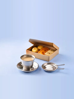 A steel tumbler of chai on a saucer beside two heaped spoons of sugar on a steel plate, with an open box of mithai behind