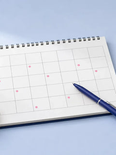 A spiral wall calendar with a few pink dots, a blue pen, a glass of water and a bowl of almonds and walnuts on a pale blue table